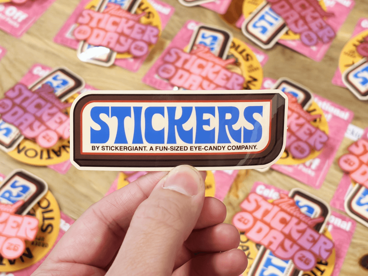 A hand holds a retro style ‘Stickers by Stickergiant’ sticker above a table covered with National Sticker Day stickers.