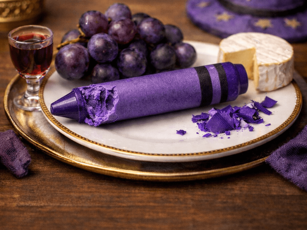 Bitten purple crayon on a plate with grapes and a wheel of cheese