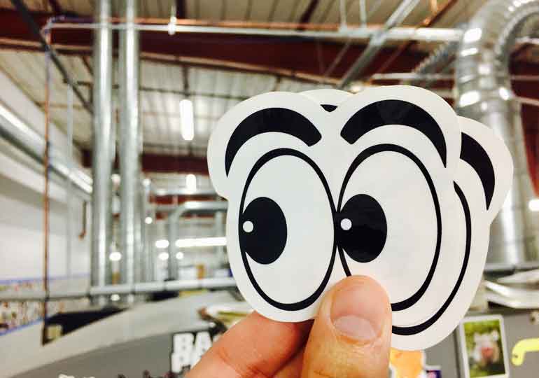 Hand holding a cartoon eyes sticker inside a production facility with pipes and equipment in the background.