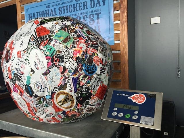 Large sticker covered ball displayed on a metal scale during an official weigh in, with the digital screen showing 231.6 pounds and a National Sticker Day banner in the background.