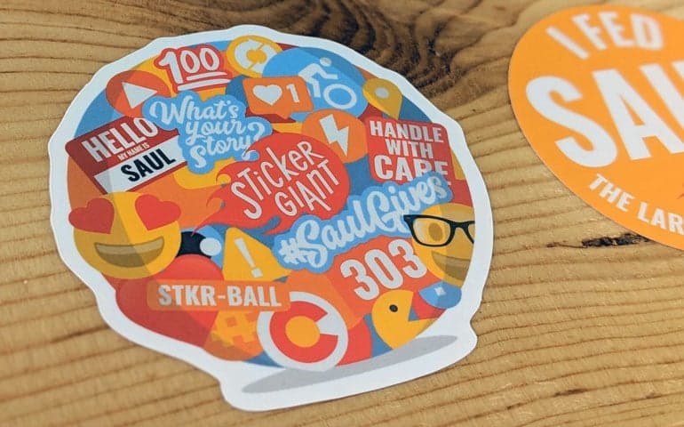 Colorful die cut sticker with Saul themed icons and StickerGiant branding on a wooden surface.