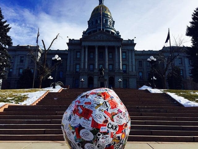 arge sticker covered ball displayed on the steps leading up to the Colorado State Capitol building, with the dome and front facade visible in the background under a blue sky.