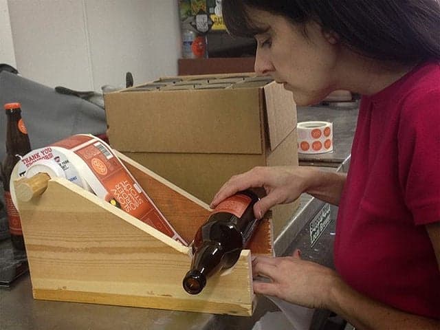 Person applying a custom label by hand to a beer bottle using a wooden labeling jig, with rolls of StickerGiant printed labels nearby.
