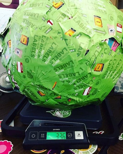 Large green sticker covered ball sitting on a digital scale, showing a weight of 7.36 pounds, with overlapping product label stickers visible on the surface.