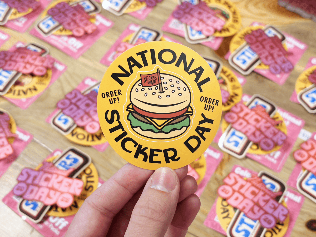 A hand holds a round yellow ‘National Sticker Day’ burger themed sticker above a table covered with similar sticker designs.