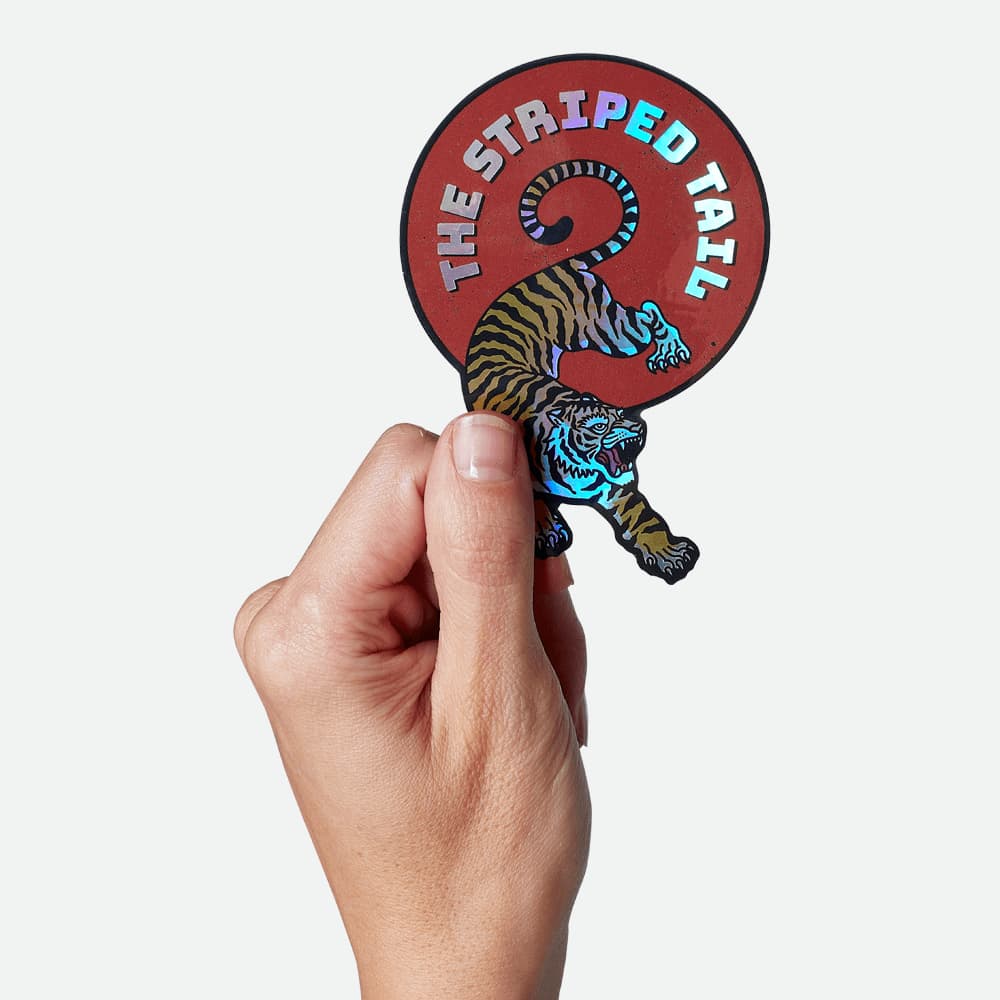 A hand holding a holographic tiger sticker that says "The Striped Tail"