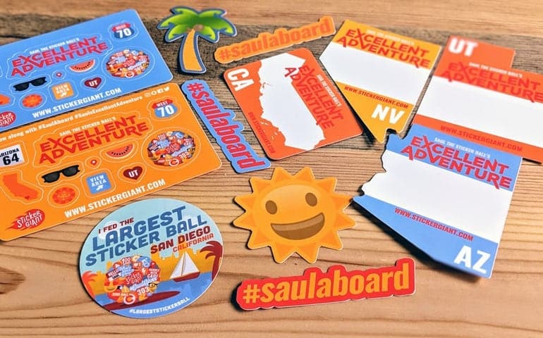 Assorted custom Excellent Adventure stickers, state shaped stickers, hashtag decals, and a round Saul sticker arranged on a wooden surface.