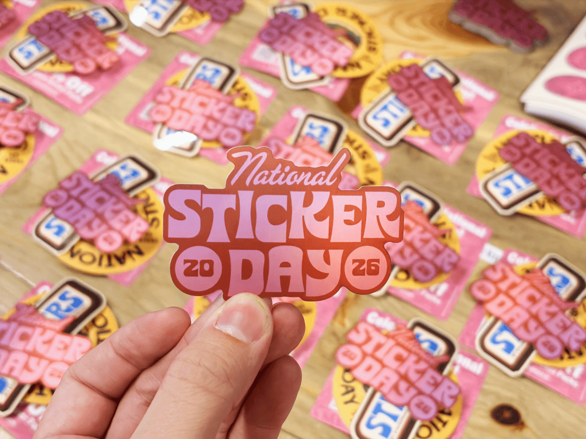 A hand holds a pink ‘National Sticker Day 2026’ sticker above a table covered with similar stickers.