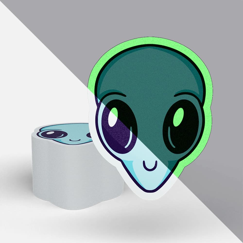 A stack of glow in the dark stickers printed by StickerGiant in the shape of an alien head - half of the picture is in daylight and half of the picture is dark showing the glow effect