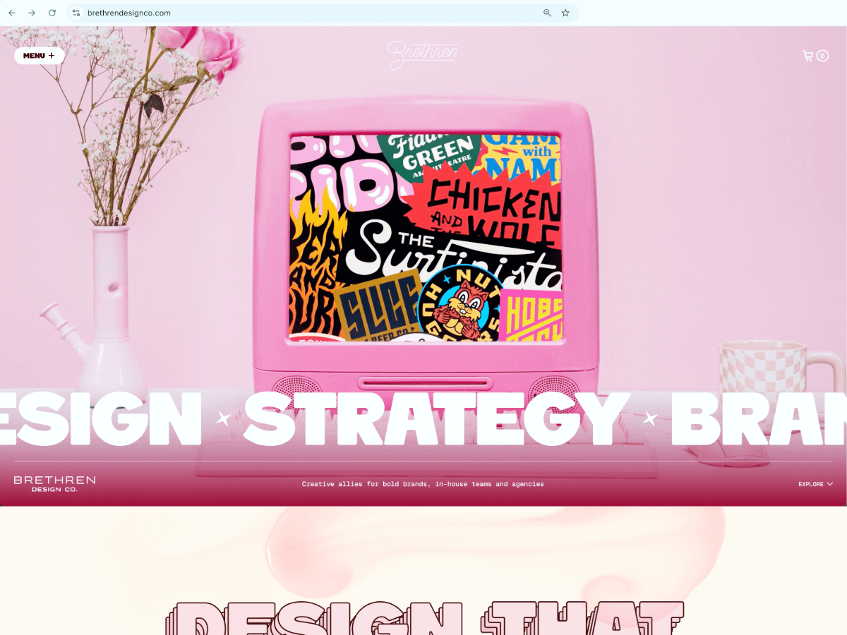 A pink retro computer displaying colorful graphic logos sits on a pink desk between a flower vase and a checkered mug on a pastel pink background.