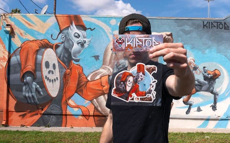 Kiptoe holding a sheet of custom Die Cut Stickers in front of one of his large mural paintings, showing how the sticker designs replicate elements of the mural artwork.